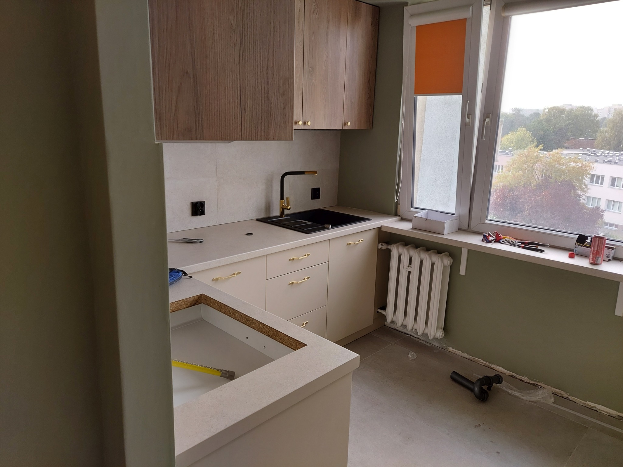 Częściowo zmontowana kuchnia z drewnianymi szafkami, czarnym zlewem z złotą armaturą i grzejnikiem pod oknem, widoczne narzędzia i elementy instalacji na podłodze.