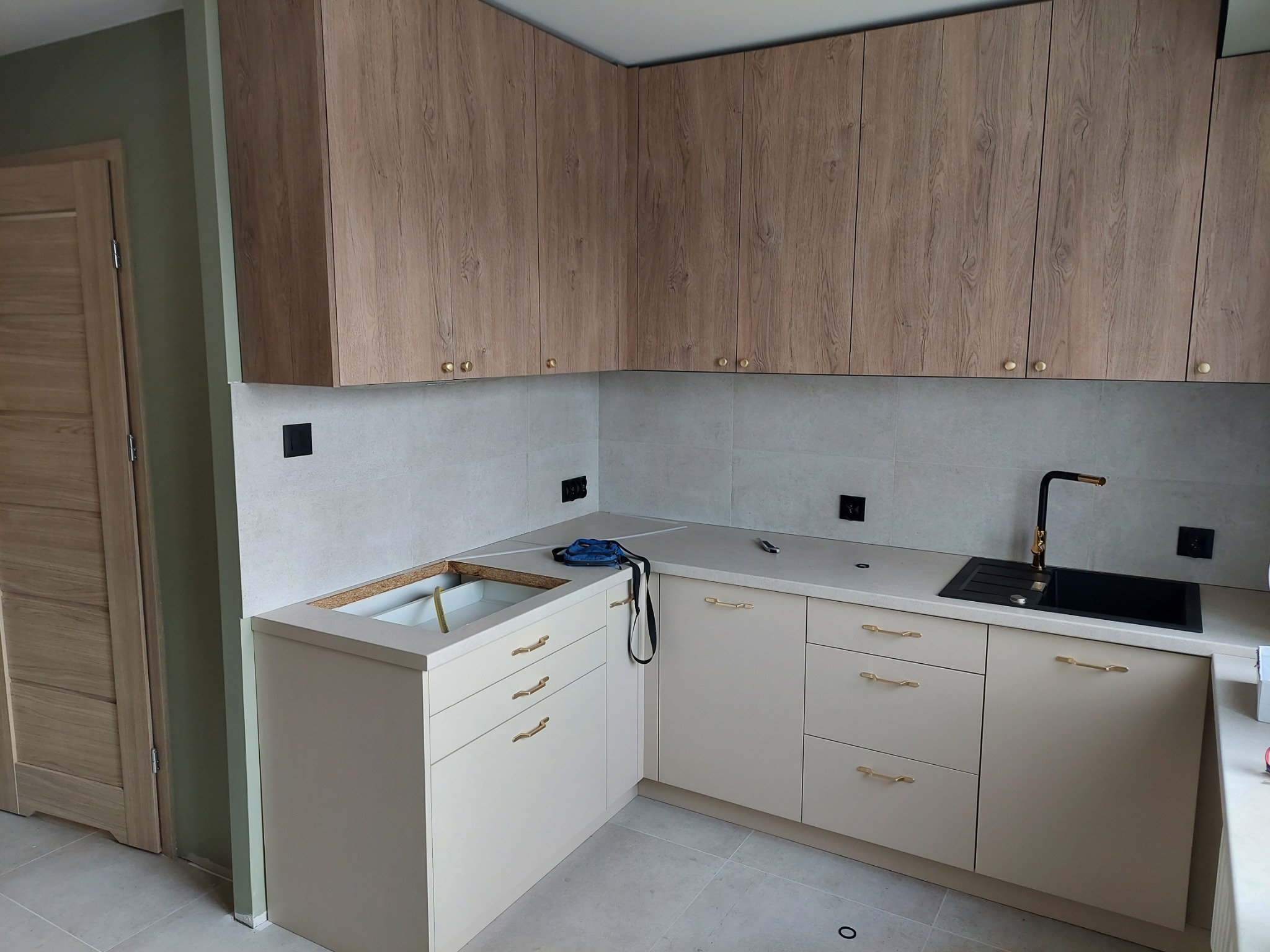 Narożna zabudowa kuchenna z jasnymi, kremowymi szafkami dolnymi z delikatnymi złotymi uchwytami i drewnopodobnymi szafkami górnymi, czarny zlew i bateria, widoczne gniazdka elektryczne na ścianie.