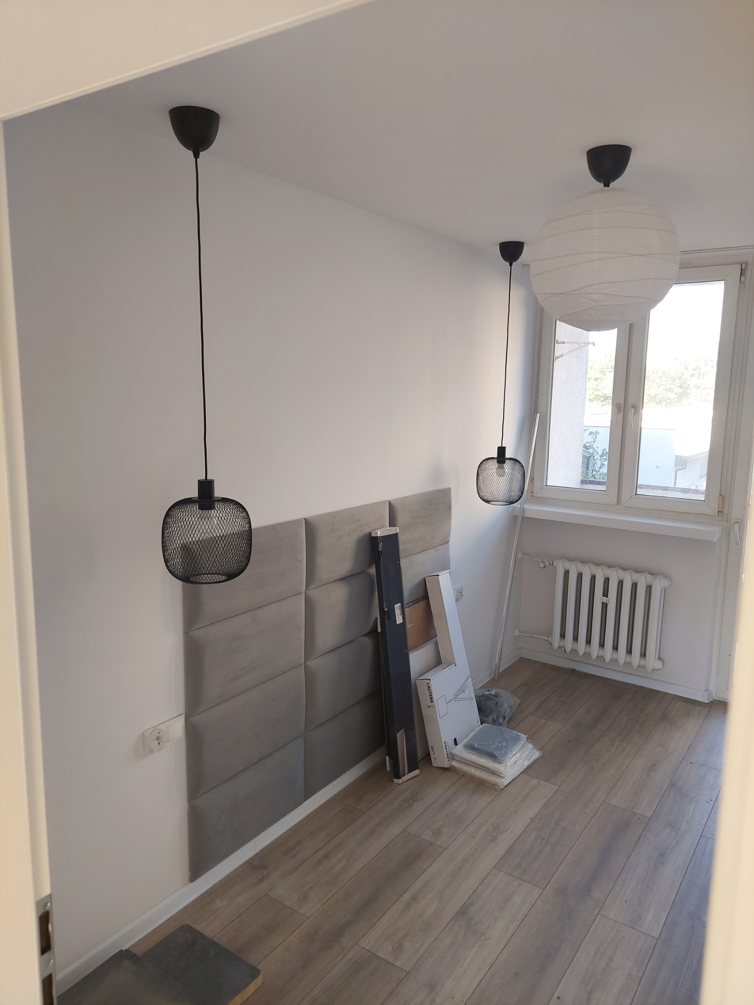 Pokój z panelami ściennymi w odcieniach szarości, drewnopodobnymi panelami podłogowymi, dwoma różnymi lampami wiszącymi i grzejnikiem pod oknem, w trakcie aranżacji.