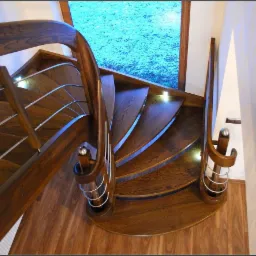 Kręcone schody z ciemnego drewna z metalowymi balustradami i podświetleniem LED, widok z góry na parkietową podłogę i okno z widokiem na zieleń.