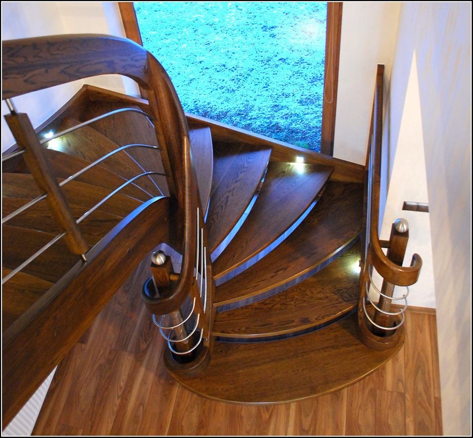 Kręcone schody z ciemnego drewna z metalowymi balustradami i podświetleniem LED, widok z góry na parkietową podłogę i okno z widokiem na zieleń.