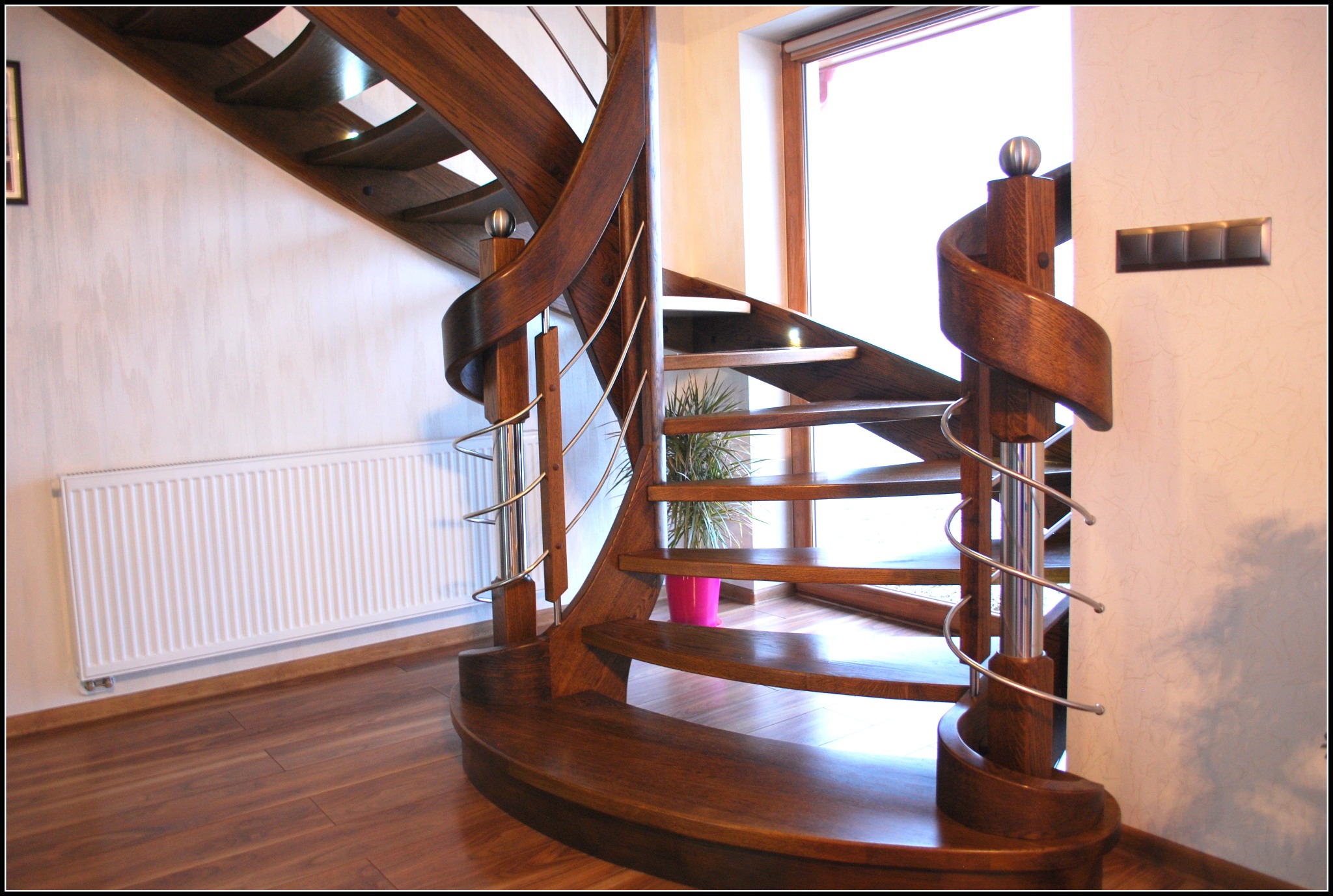 Drewniane schody spiralne z ciemnego drewna z metalowymi poręczami, widok z dołu, w tle okno i roślina w różowej doniczce, na ścianie biały grzejnik.