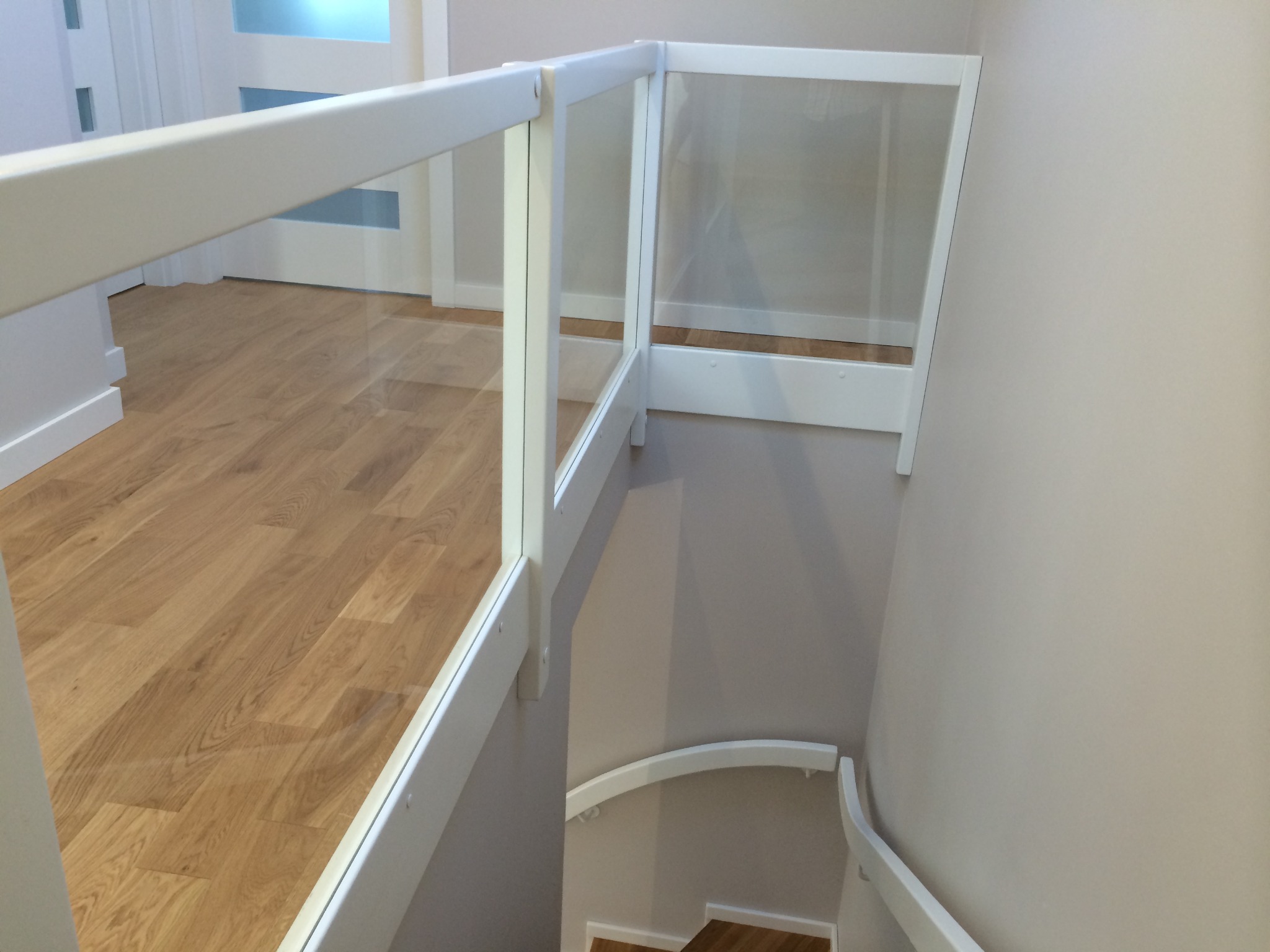 Białe schody z drewnianymi stopniami i szklaną balustradą, widok z góry na dół, z widocznymi fragmentami podłogi i drzwi w tle.