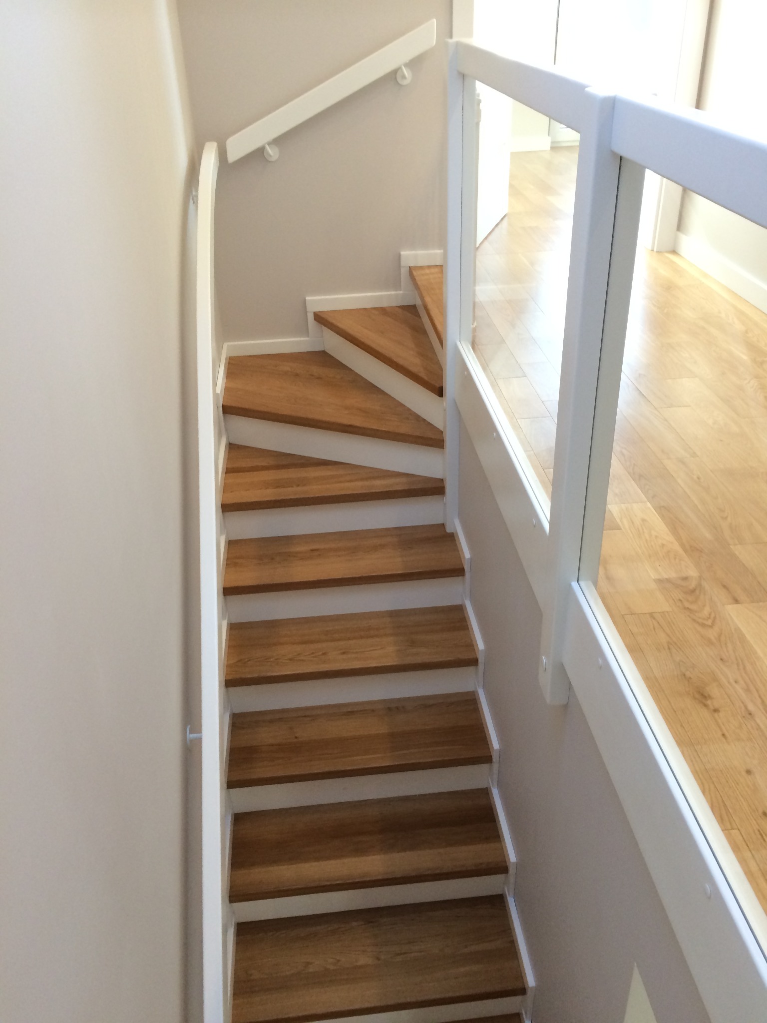 Klatka schodowa z drewnianymi stopniami i białymi podstopnicami, balustrada z białych słupków i przezroczystego szkła, widok z góry na parkiet na piętrze.
