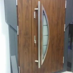 Brązowe drzwi zewnętrzne z przeszkleniem i srebrnym uchwytem, prezentowane w salonie w Łodzi, z widocznym logo producenta 'Marton' nad drzwiami.