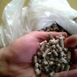 Garść pelletu drzewnego na dłoni, z workiem wypełnionym pelletem w tle. Widoczna struktura i kolor pelletu. Naturalne światło.