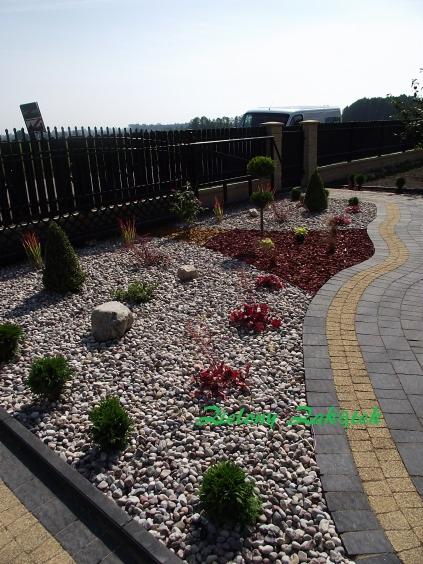 Aranżacja ogrodu z jasnymi kamieniami, czerwonymi rabatami z kory, niskimi drzewkami i krzewami, oraz ścieżką z kostki brukowej w odcieniach szarości i żółci, ogrodzona czarnym metalowym płotem.