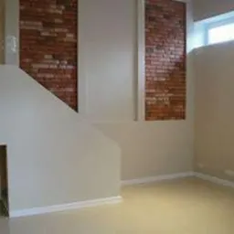 Wnętrze pokoju po remoncie z fragmentem ściany z cegły klinkierowej, pomalowane ściany w odcieniach beżu i jasny parkiet. Widoczne okno w górnej części kadru.