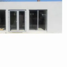 Elewacja budynku z trzema szklanymi elementami: dwuskrzydłowe drzwi pośrodku, okno po lewej i prawej stronie. Jasna kolorystyka. Widoczne narzędzia wewnątrz budynku.