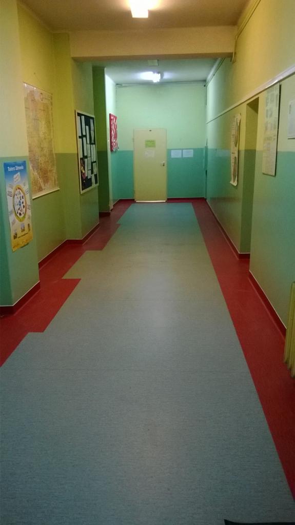 Długi korytarz z szarą wykładziną i czerwonymi listwami przypodłogowymi. Na ścianach wiszą plakaty i tablice informacyjne. Widok w perspektywie zbieżnej.