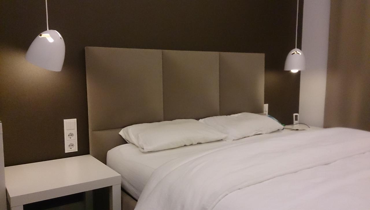 Eleganckie łóżko z tapicerowanym wezgłowiem w odcieniach beżu, oświetlone lampami wiszącymi. Stonowana kolorystyka ścian. Widoczny stolik nocny i gniazdka elektryczne.