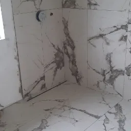 Wykończona łazienka z białymi płytkami imitującymi marmur, widoczne przyłącza wodne i odpływ liniowy w podłodze.