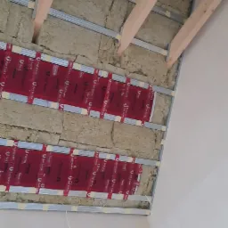 Izolacja poddasza z widocznymi belkami drewnianymi, metalowymi profilami i czerwoną folią izolacyjną Red Snake, zamocowaną taśmą. Widok z dołu na strop przed montażem płyt gipsowo-kartonowych.