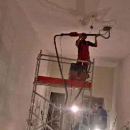 Pracownik na rusztowaniu szlifuje sufit szlifierką z odkurzaczem, przygotowując powierzchnię pod malowanie. Widoczne ślady po szpachlowaniu.