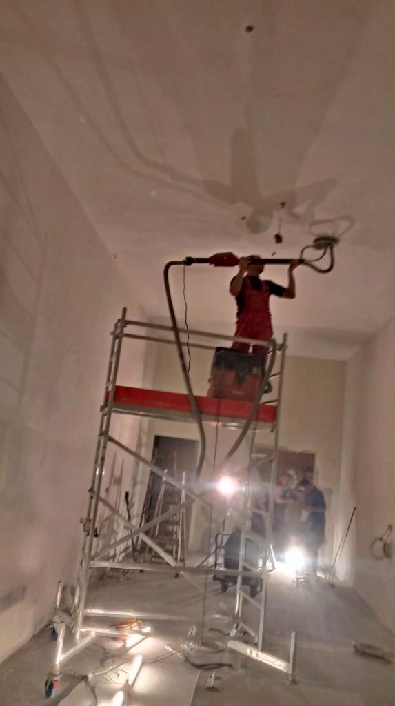Pracownik na rusztowaniu szlifuje sufit szlifierką z odkurzaczem, przygotowując powierzchnię pod malowanie. Widoczne ślady po szpachlowaniu.