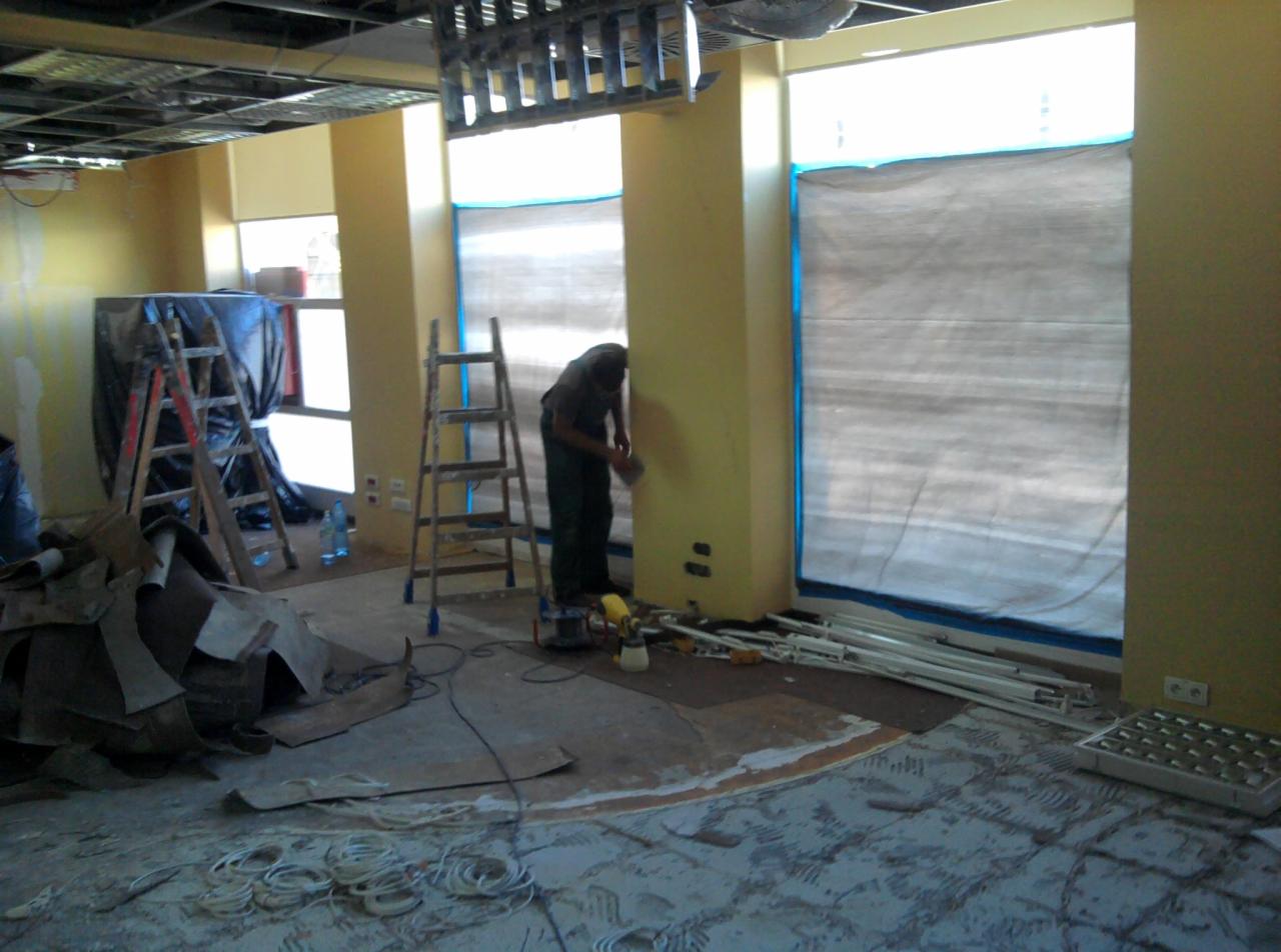 Prace wykończeniowe wnętrza: szlifowanie ściany w żółtym pomieszczeniu z osłoniętymi oknami, widoczne drabiny, narzędzia i przygotowane materiały.