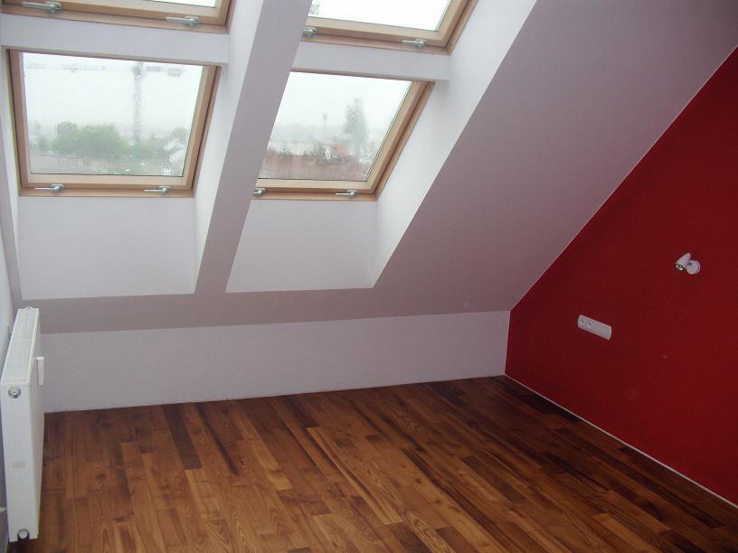 Poddasze po remoncie: drewniana podłoga, białe ściany, czerwona ściana akcentująca, okna dachowe w skosach. Grzejnik przy ścianie.