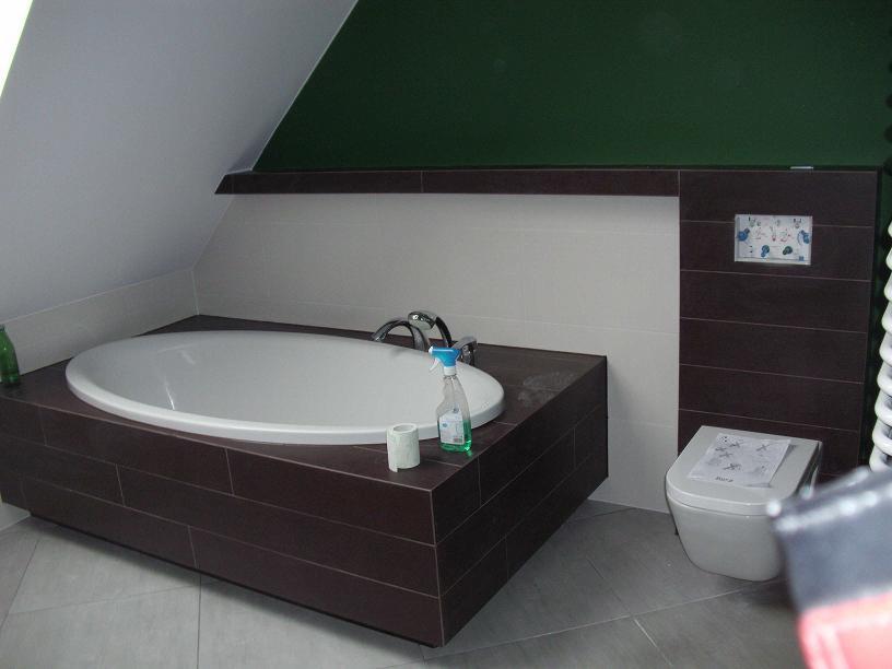 Wykończona łazienka na poddaszu: owalna wanna obudowana brązowymi płytkami, toaleta z instrukcją, białe i brązowe płytki na ścianach, zielona ściana pod skosem.