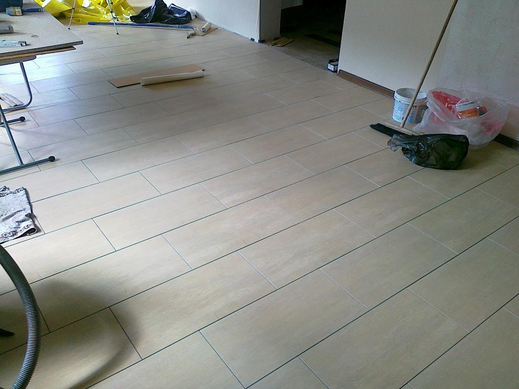 Ułożona podłoga z płytek w odcieniach beżu, z widocznymi fugami i elementami wyposażenia wnętrza w tle. Widok z góry, ukośne ułożenie płytek.