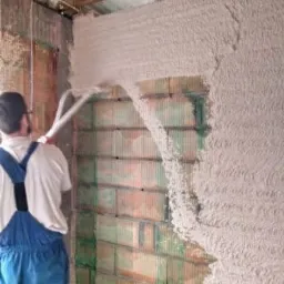 Tynkarz w niebieskim kombinezonie nanosi tynk maszynowy na ścianę z czerwonej cegły, widoczne drewniane elementy konstrukcji dachu.