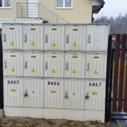 Zestaw skrzynek pomiarowych z numerami 8465, 8466, 8467, zabezpieczonych ogrodzeniem drewnianym i metalowym. Widoczny napis 'POMIAR' i znak ostrzegawczy.