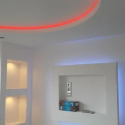 Białe ściany z wnękami, podświetlane LED: czerwone na suficie, niebieskie i żółte we wnękach. W jednej wnęce gniazdka i pudełka. Minimalistyczny design wnętrza.