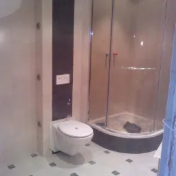 Nowoczesna łazienka z kabiną prysznicową i podwieszaną toaletą. Szare płytki podłogowe z ciemnymi akcentami. Ciemny pas płytek na ścianie.