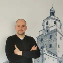 Mężczyzna z założonymi rękami na tle ściany z rysunkiem wieży zegarowej.