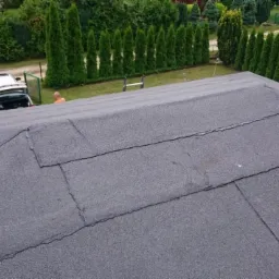 Widok z góry na dach pokryty papą, z widocznymi łączeniami i drobnymi uszkodzeniami, w tle zieleń drzew i krzewów, drabina oparta o dach, a w oddali zaparkowany samochód.