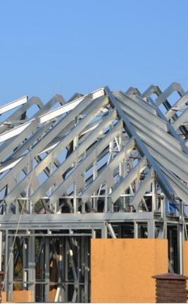 Konstrukcja domu szkieletowego w trakcie budowy, stalowe profile tworzące szkielet dachu na tle błękitnego nieba. Widoczny fragment ściany z płyt.