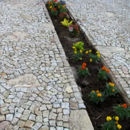Ścieżka z kostki granitowej z rabatą kwiatową pośrodku, ułożona z drobnej kostki, widok z góry, ciepłe barwy kwiatów kontrastują z szarością kamienia.