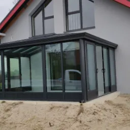 Szklana konstrukcja dobudowana do domu z szarymi ścianami i czerwonymi elementami dachu, widoczny samochód wewnątrz, teren wokół w trakcie prac ziemnych.