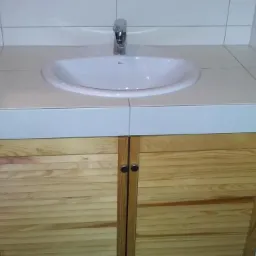 Biała umywalka wbudowana w blat z płytek, szafka z drewnianymi lamelami. Ukończona aranżacja łazienki z widocznymi detalami wykończenia.