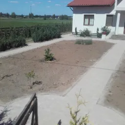 Widok z góry na posesję z nowo ułożonym chodnikiem z kostki brukowej, przygotowaną ziemią pod trawnik i nasadzenia, oraz białym domem z czerwonym dachem w tle.