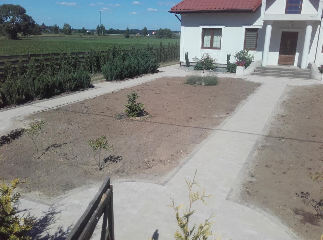 Widok z góry na posesję z nowo ułożonym chodnikiem z kostki brukowej, przygotowaną ziemią pod trawnik i nasadzenia, oraz białym domem z czerwonym dachem w tle.