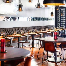 Elegancka restauracja z drewnianą podłogą, rzędami stolików nakrytych kieliszkami, ciemnobrązową tapicerowaną ławą wzdłuż ściany i designerskimi lampami wiszącymi o metalowej konstrukcji.