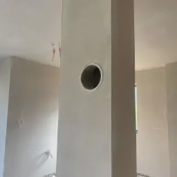 Wnętrze po nałożeniu gładzi gipsowej z widocznym otworem wentylacyjnym w kolumnie, przewody elektryczne wystające ze ściany, okno z widokiem na drzewa.