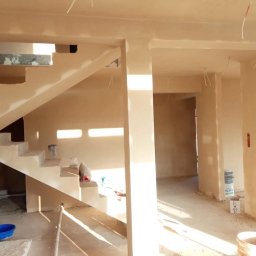 DARBUD DARIUSZ STAŃCZYK - Wnętrze domu w trakcie prac tynkarskich, widoczne otynkowane ściany, schody z częściowo położonym tynkiem oraz puste pojemniki po materiałach budowlanych.