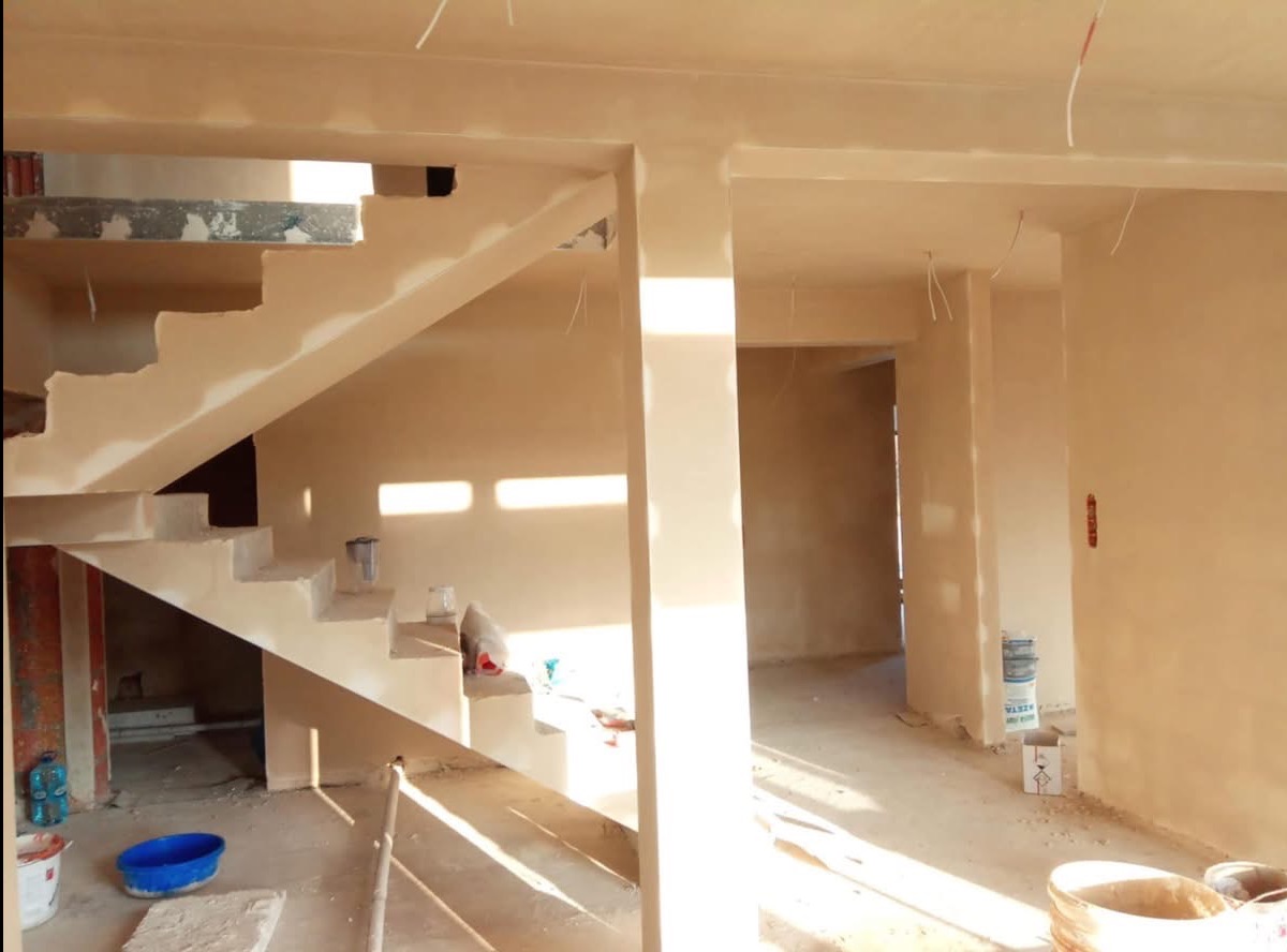 Wnętrze domu w trakcie prac tynkarskich, widoczne otynkowane ściany, schody z częściowo położonym tynkiem oraz puste pojemniki po materiałach budowlanych.