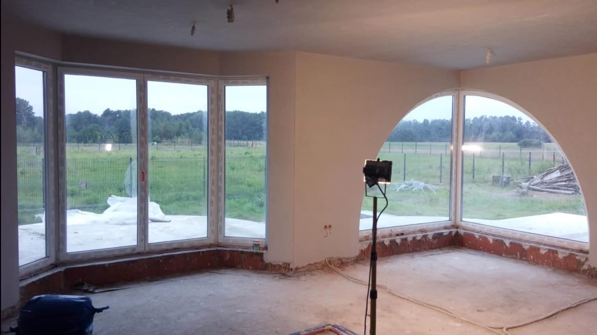 Wnętrze w trakcie prac wykończeniowych: świeżo otynkowane ściany, duże okna z widokiem na zewnątrz, widoczne przewody elektryczne przy podłodze, lampa budowlana na statywie.