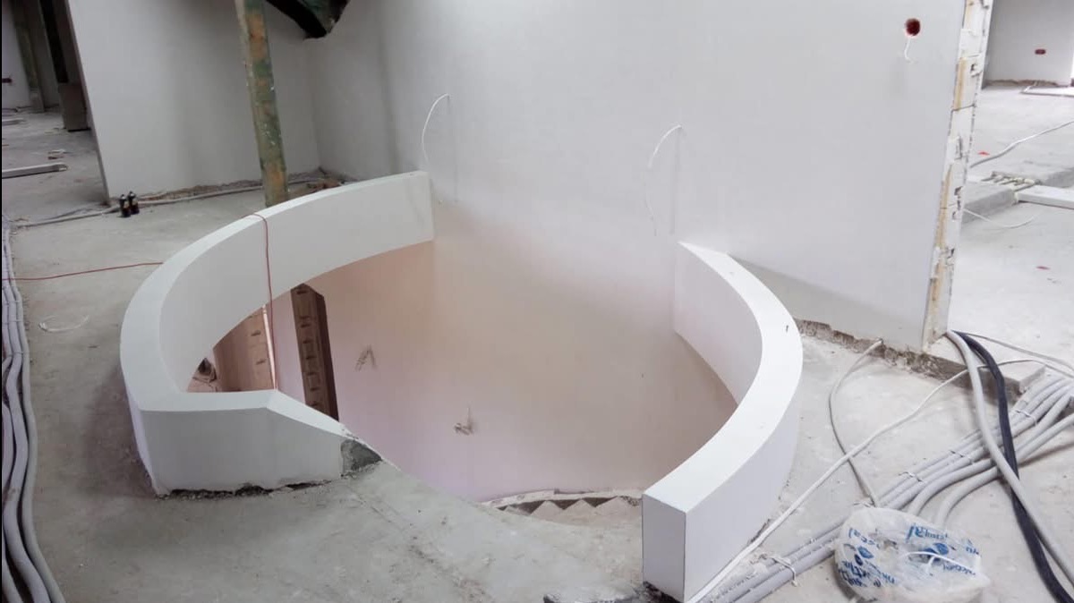 Wnętrze w trakcie remontu z widoczną, częściowo wykończoną, spiralną konstrukcją schodów z białego tynku, surowy beton na podłodze i wystające kable elektryczne.