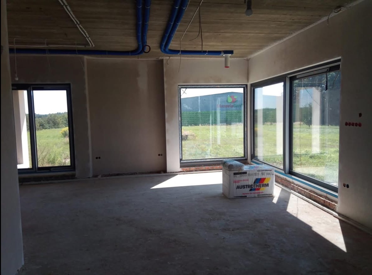 Surowe wnętrze z tynkami na ścianach, widoczne instalacje na suficie i paczka styropianu Austrotherm przy oknie.