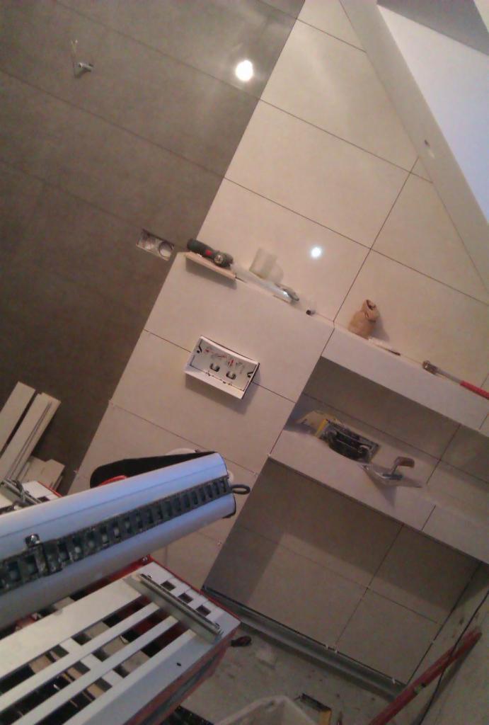 Ściana wyłożona płytkami w trakcie prac. Widoczna maszynka do cięcia płytek, narzędzia glazurnicze, puszka elektryczna. Częściowo ułożone płytki w odcieniach szarości i beżu.