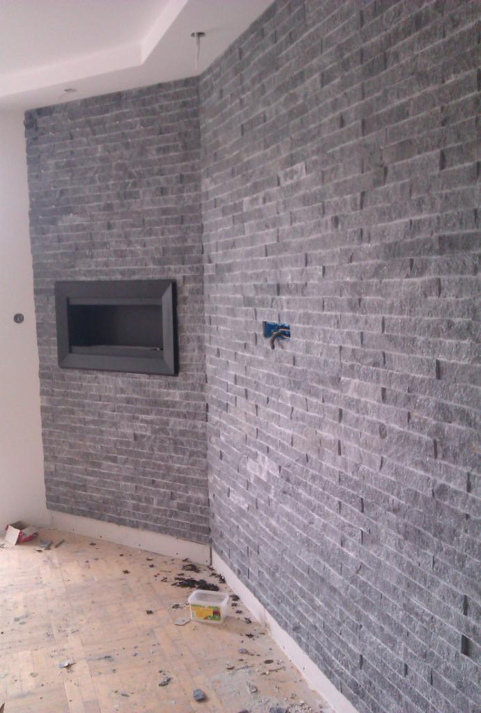 Ściana wyłożona szarą, wąską cegłą dekoracyjną. W ścianie widoczna wnęka na kominek i wystające kable elektryczne. Na podłodze widoczne ślady po pracy.