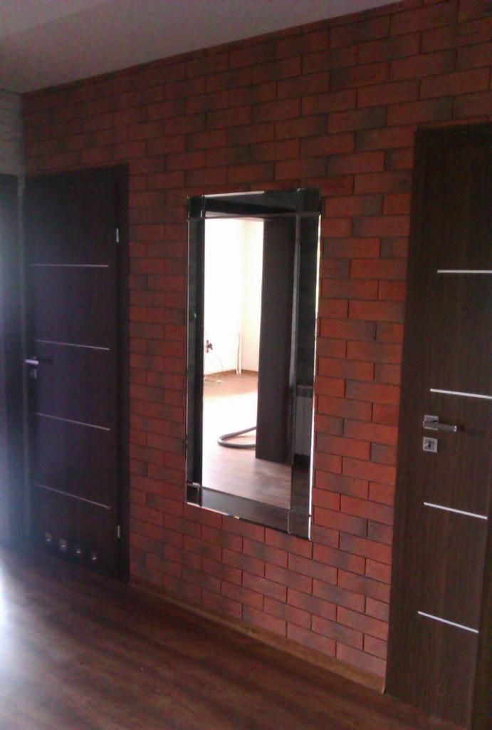 Ściana z cegły klinkierowej z lustrem w ramie, dwie ciemne, drewniane drzwi z poziomymi zdobieniami. Podłoga z paneli w odcieniach brązu. Wnętrze mieszkania.