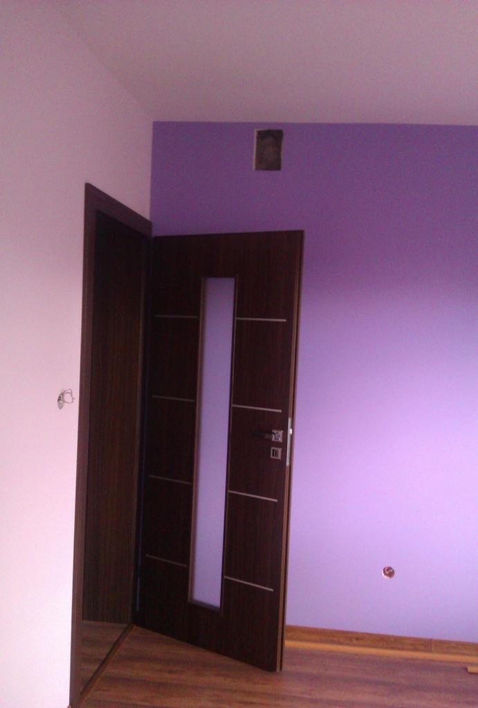 Ciemne, drewniane drzwi z przeszkleniem w otwartym ościeżu, na tle fioletowej i różowej ściany. Widoczne fragmenty podłogi z paneli.