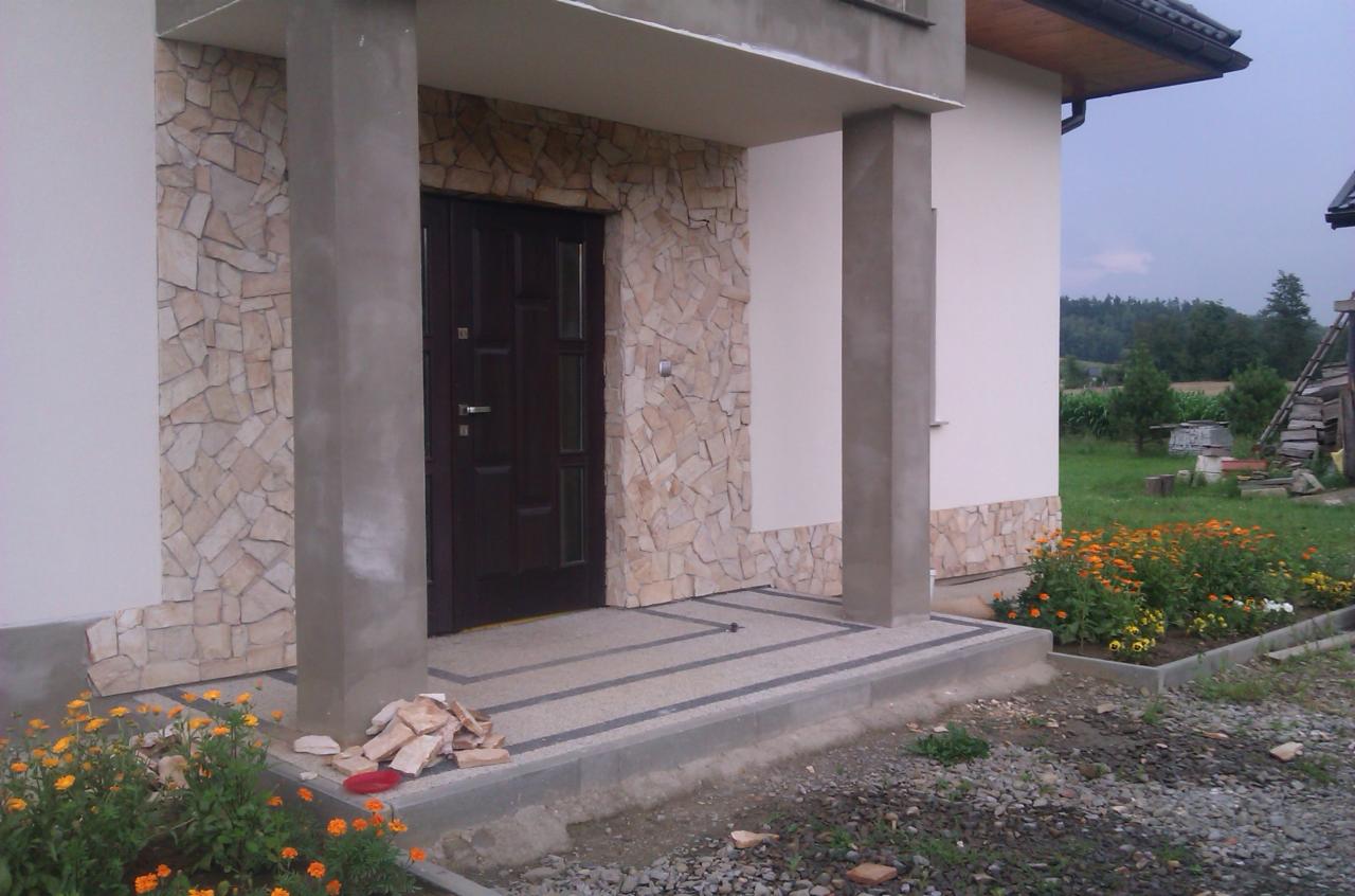 Wykończone wejście do domu z kamienną okładziną ścian i kolumn. Na podłożu widoczne płytki i kamienie. W tle zieleń i elementy krajobrazu.