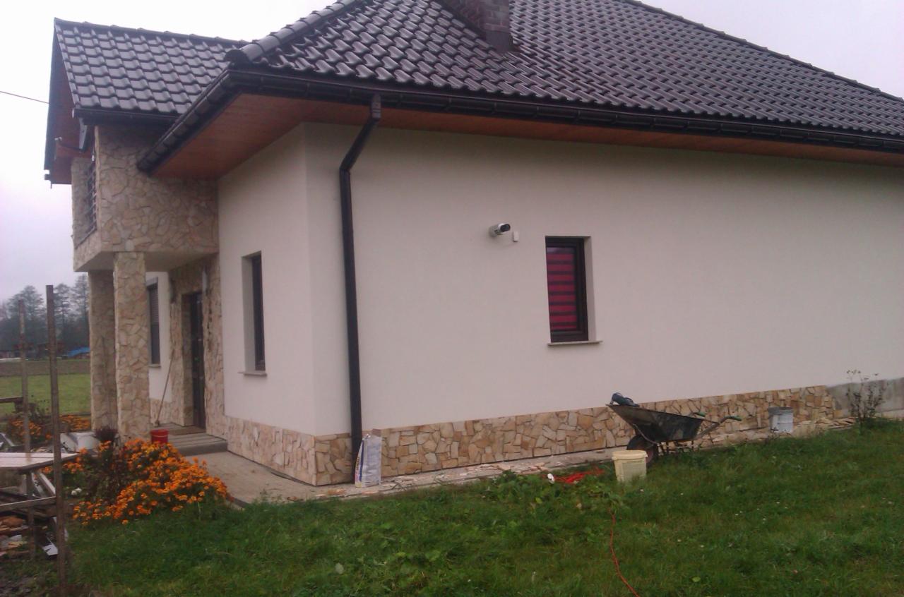 Elewacja domu jednorodzinnego z elementami kamiennymi i brązową dachówką. Widoczny fragment ogrodu i taczka. Ujęcie z boku budynku.