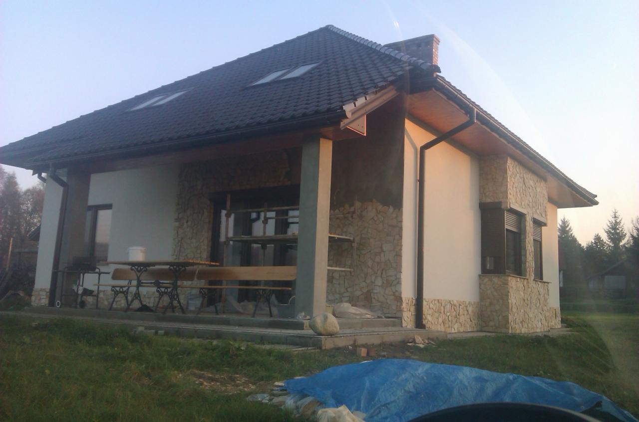 Dom jednorodzinny w trakcie wykończenia elewacji kamiennej, widoczne rusztowania i materiały budowlane. Dach pokryty dachówką, okna z roletami zewnętrznymi.