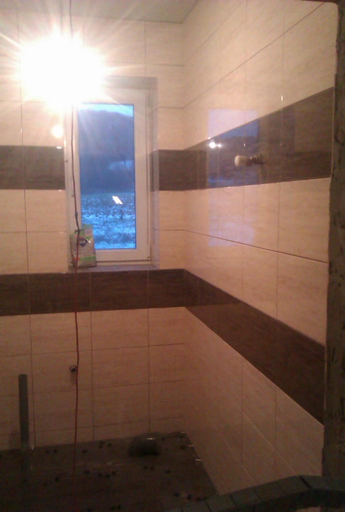 Wykończona łazienka z beżowymi i brązowymi płytkami, widok na okno z zewnątrz. Na parapecie stoi opakowanie. Widoczny fragment ściany w stanie surowym.